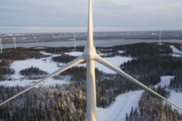 Blick auf eine winterliche skandinavische Waldlandschaft. Im Vordergrund befindet sich ein Windrad. Im Hintergrund sind weitere Windräder sowie ein Fluss zu erkennen.