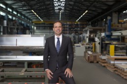 Man in antracite suit stands frontal and picture center with confident smile in a factory hall