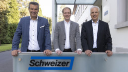 Drei Männer im Anzug stehen hinter einer hüfthohen wand mit dem Logo von der Ernst Schweizer AG