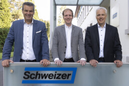 Drei Männer im Anzug stehen hinter einer hüfthohen wand mit dem Logo von der Ernst Schweizer AG