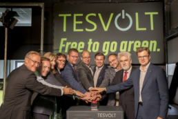 Eine Menschengruppe steht gemeinsam vor einem großen TESVOLT-Schild und legt die Hände übereinander. Im Hintergrund steht der Schriftzug ‚TESVOLT – Free to go green.